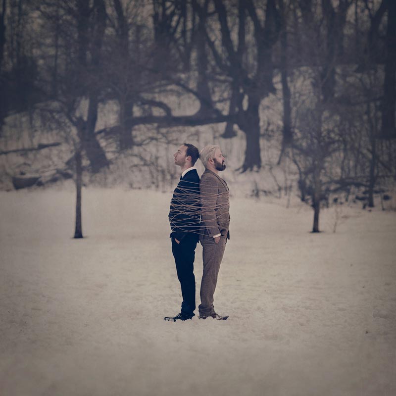 Two men stand back to back, tied together in snowy field