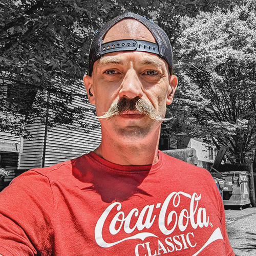 A photo of a man with a backwards black cap and a large mustache, wearing a coca-cola t-shirt